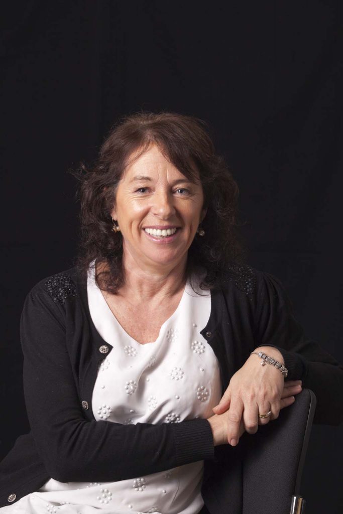 Olive O'Grady is sitting in front of a black background, wearing a white top and black cardigan. She is smiling and looking towards the camera.